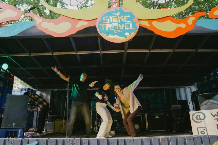 Group of friends dancing on stage under a colourful “Stoke Travel Co.” sign, drinks in hand, during a live music set at the Munich Oktoberfest campsite.