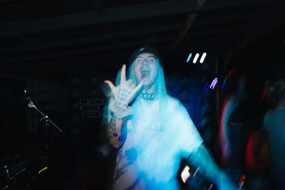 Performer at the Oktoberfest campsite party throwing a hand sign under blue stage lights, surrounded by a dancing crowd and electric night energy.