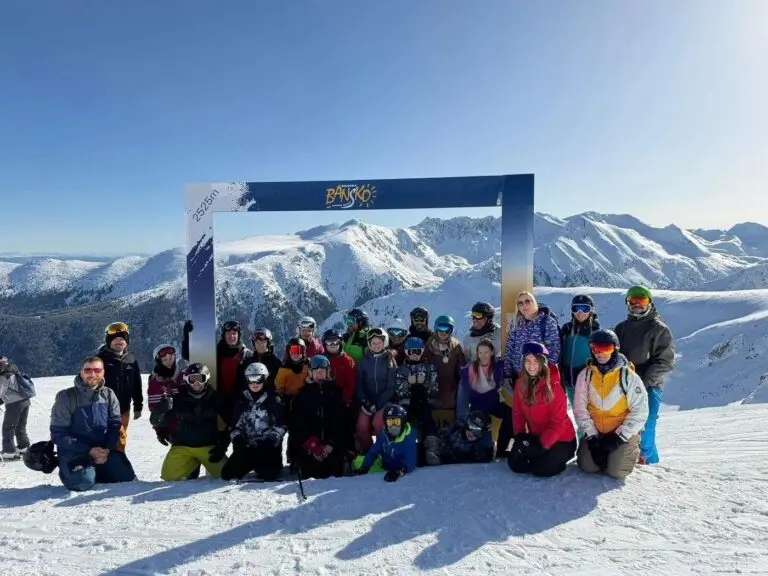 Large group of skiers and snowboarders posed under a summit archway on a Bansko mountaintop, snowy Pirin peaks behind