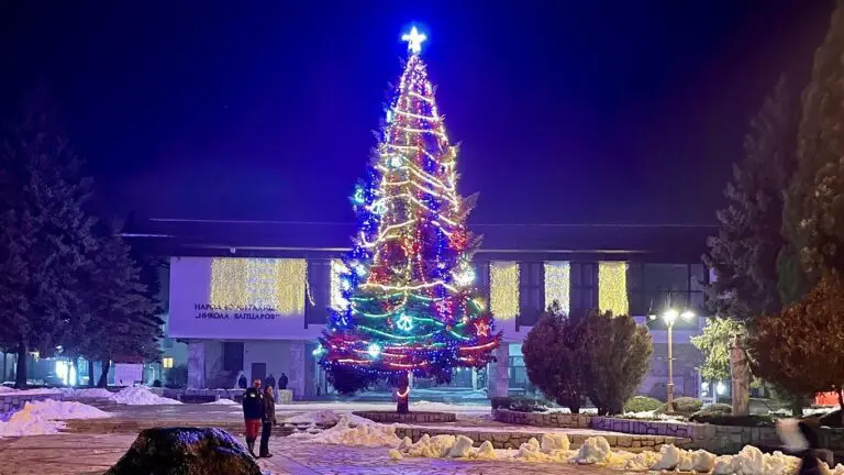 A huge Christmas tree covered in coloured lights and a glowing star lit up at night in Bansko's snowy town square, with festive lights strung across the building behind it.