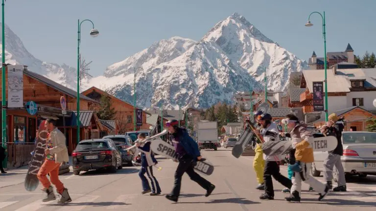 Skiers crossing a sunny resort street in Les Deux Alpes with a snow-covered peak behind