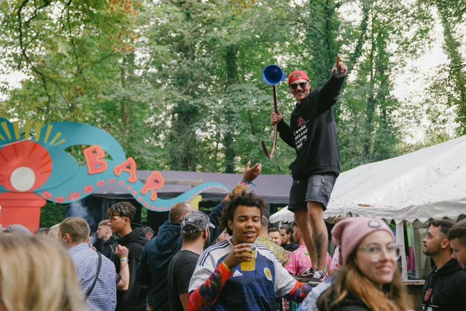 Festivalgoers enjoy Oktoberfest celebration at an outdoor beer garden, with a man holding a beer bong and pointing excitedly above a lively crowd near a colorful bar sign surrounded by trees.