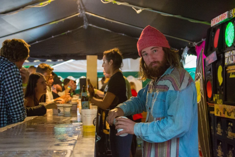 Bartender in a beanie serves drinks at a busy Oktoberfest bar tent as festivalgoers line up for beer and cocktails under ambient lighting